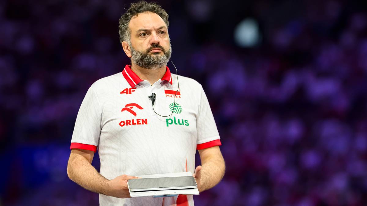 Mężczyzna w białej koszulce z logo "ORLEN PLUS" trzymający tablicę, z rozmazanym tłem widowni.