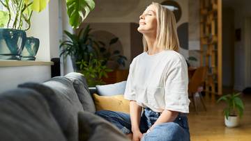 Kobieta siedzi na kanapie z głową uniesioną w kierunku światła, symbolizując wdzięczność lub spok&oacute;j.