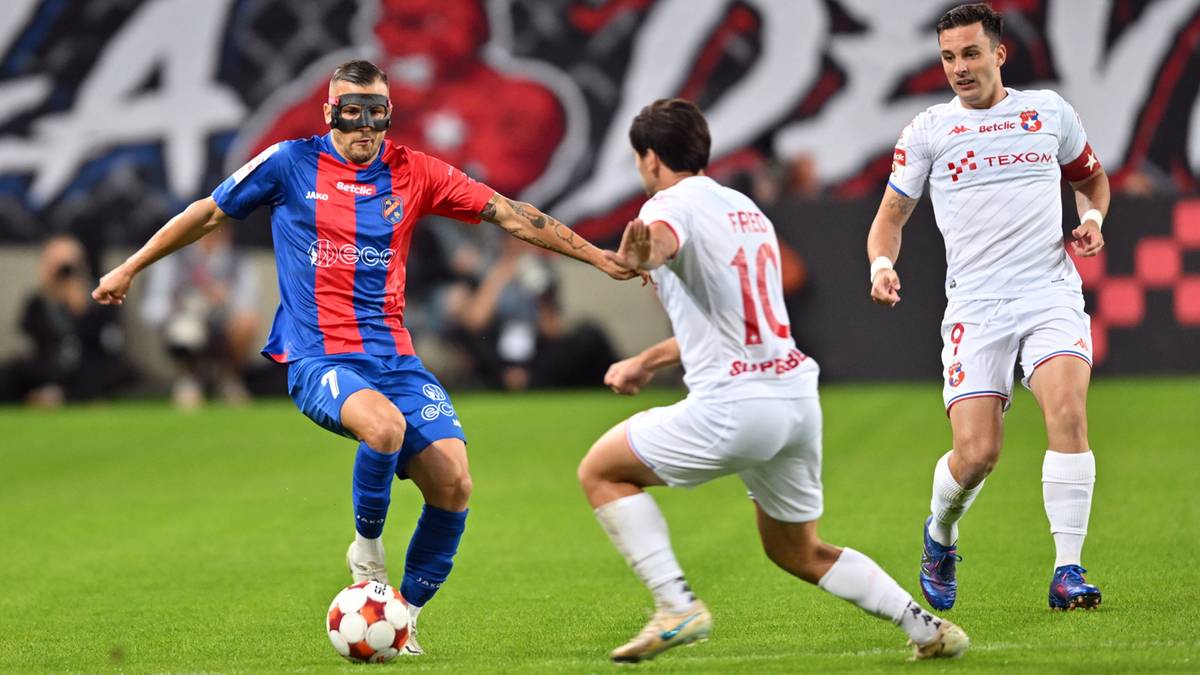 Piłkarz w niebiesko-czerwonym stroju z maską na twarzy drybluje piłką, podczas gdy dwóch zawodników w białych strojach próbuje mu ją odebrać na zielonym boisku.