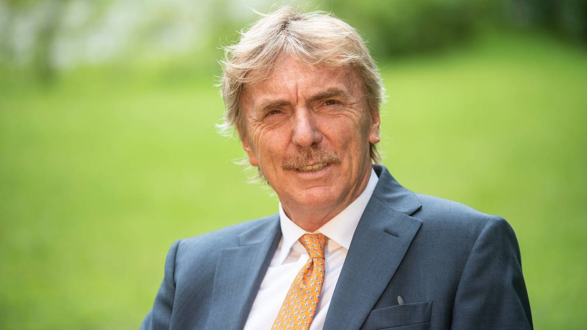 Mężczyzna w średnim wieku o blond włosach i wąsach, ubrany w ciemny garnitur i pomarańczowy krawat, uśmiecha się do aparatu.