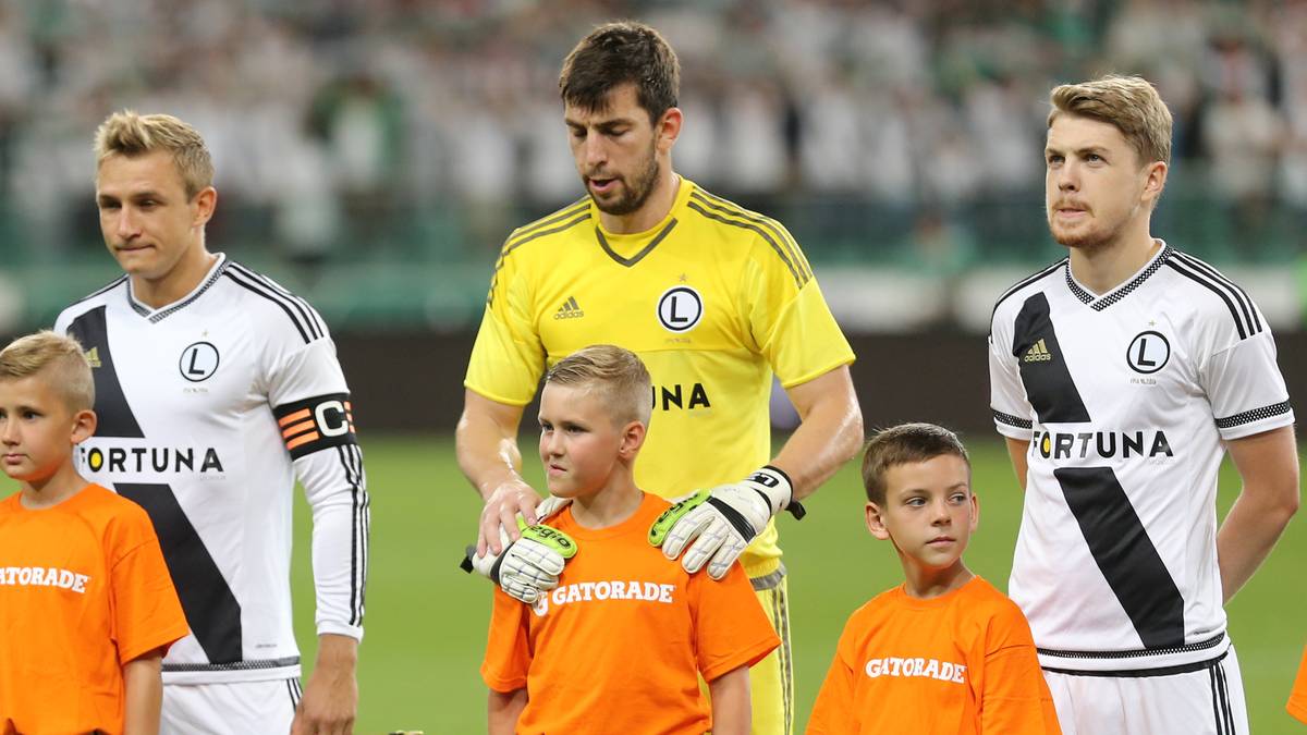 Trzech dorosłych piłkarzy i dwóch młodych chłopców stoi na zielonym boisku piłkarskim.