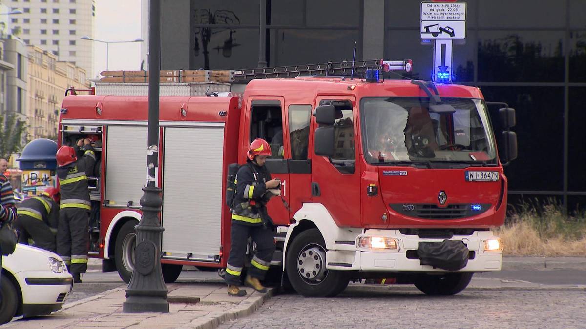 Samochód strażacki z włączonymi sygnałami świetlnymi, obok niego strażacy w akcji.