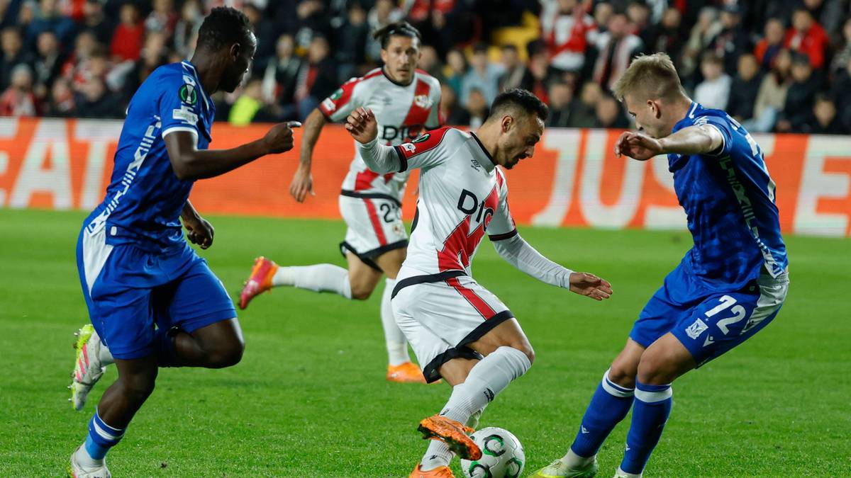 Trzech piłkarzy na boisku podczas meczu piłki nożnej, dwaj w niebieskich strojach i jeden w białej koszulce z czerwonymi elementami, walczą o piłkę.