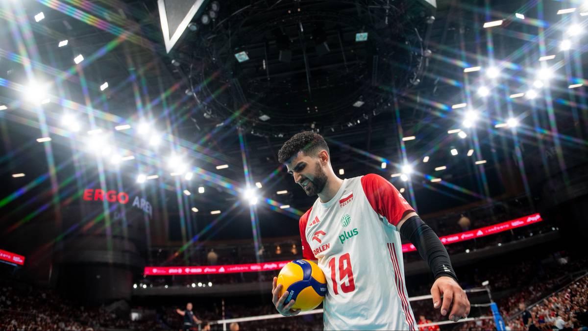 Siatkarz w koszulce z numerem 19, trzymający piłkę do siatkówki, stoi na tle oświetlonej hali sportowej.