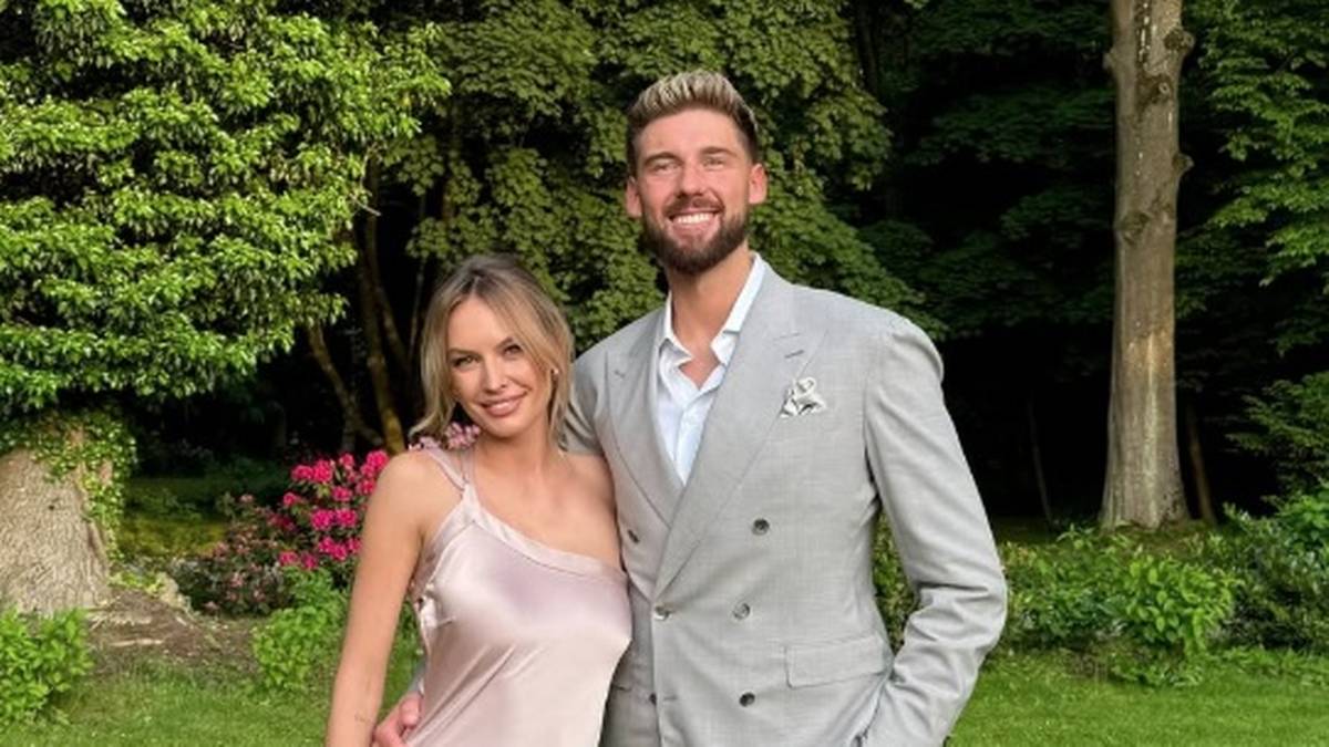 Uśmiechnięta para pozuje do zdjęcia na tle zieleni, mężczyzna w garniturze, kobieta w sukience.