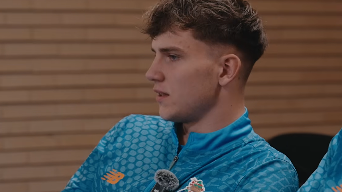 Portret młodego mężczyzny w sportowej bluzie, siedzącego i mówiącego do mikrofonu.