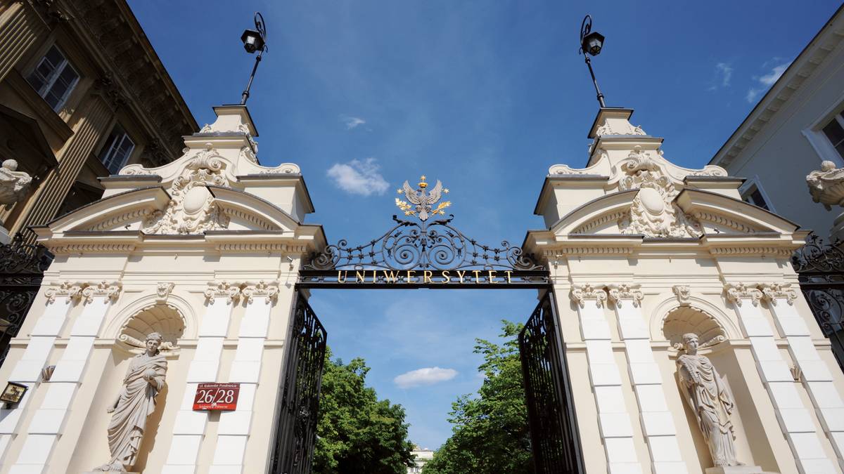 Monumentalna brama Uniwersytetu Warszawskiego z ozdobnymi elementami architektonicznymi i zieloną aleją w tle.