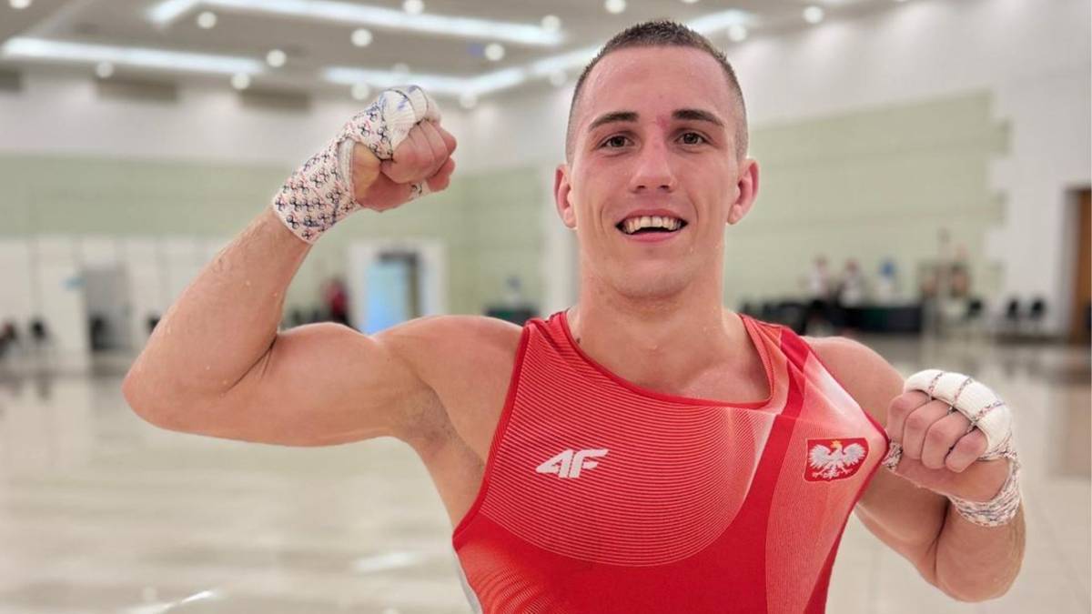 Pięściarz w czerwonym stroju sportowym z biało-czerwoną naszywką z orłem, prezentuje swoje mięśnie bicepsów, zaciskając pięści.