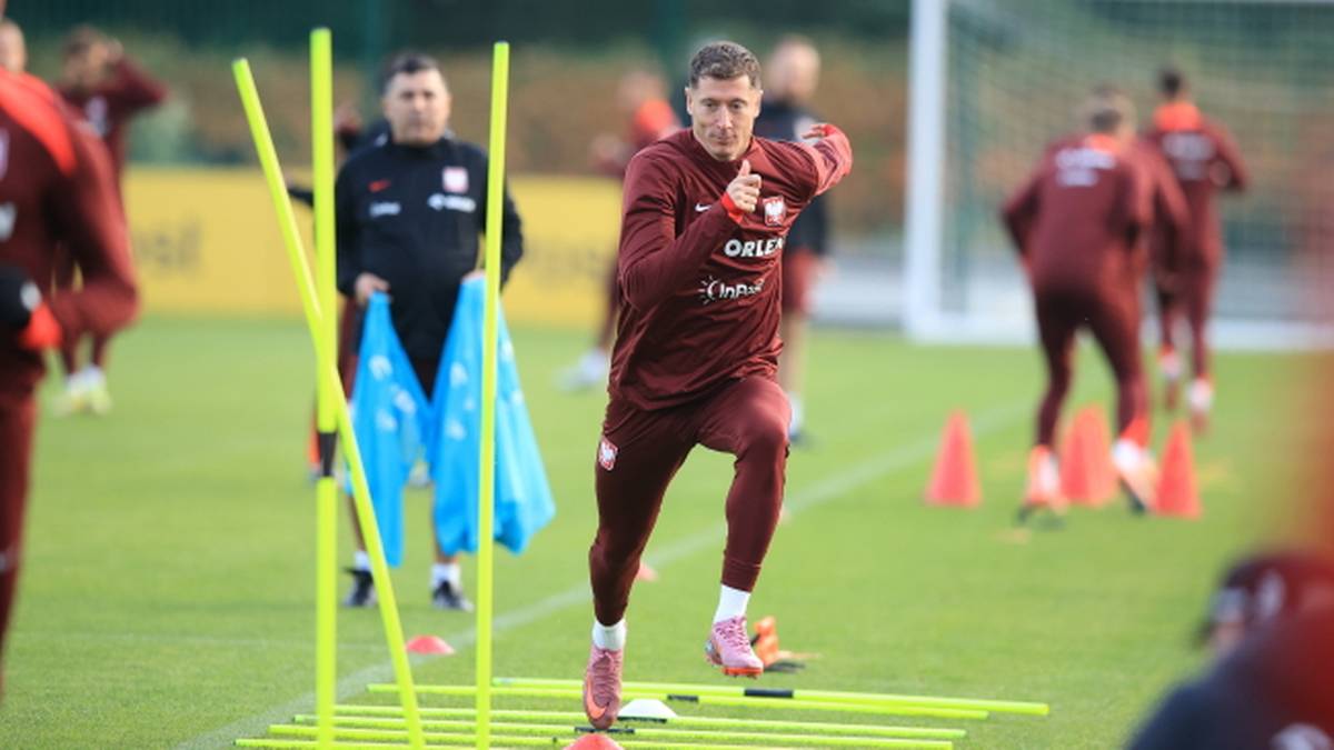 Piłkarz Robert Lewandowski w koszulce reprezentacji Polski trenuje bieganie między żółtymi tyczkami na boisku.