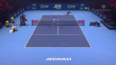 Dw&oacute;ch zawodnik&oacute;w grających w tenisa na niebieskim korcie, jeden z nich po lewej stronie siatki, drugi po prawej. W tle widać bandy reklamowe i trybuny.