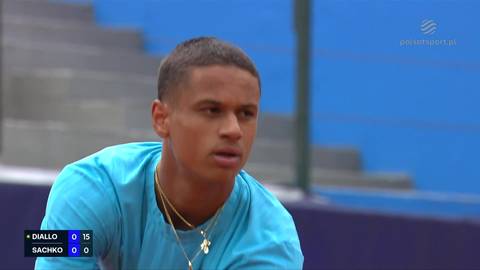 Młody mężczyzna o ciemnej karnacji w niebieskiej koszulce z dwoma złotymi naszyjnikami na szyi patrzy w bok. Na dole po lewej stronie widnieje tablica wynik&oacute;w meczu tenisowego.