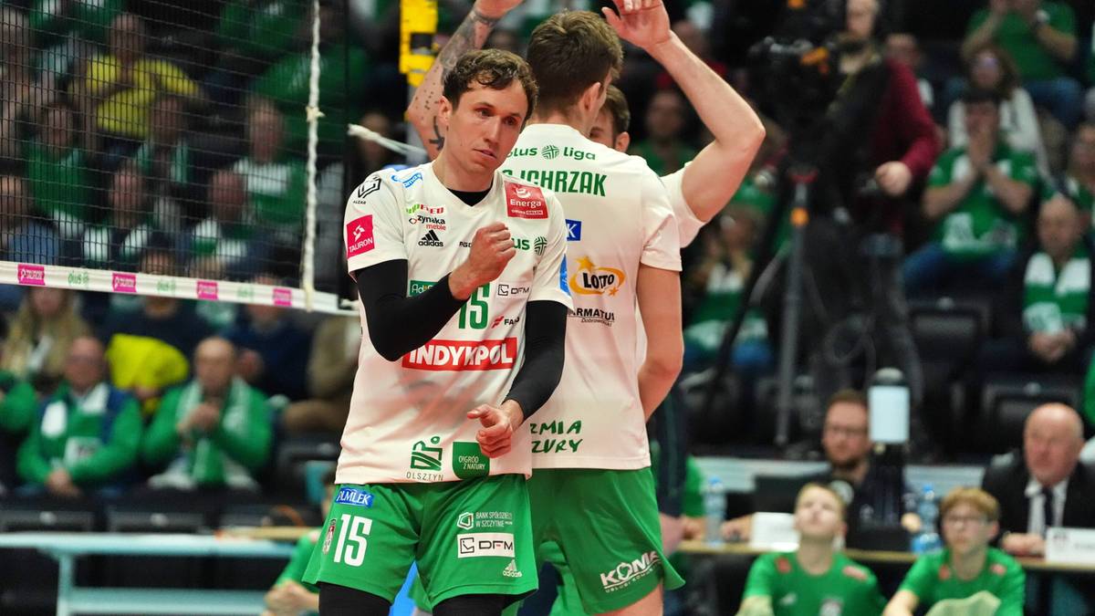 Siatkarze Indykpol AZS Olsztyn w białych koszulkach i zielonych spodenkach stoją na boisku siatkarskim, patrząc w stronę przeciwną do obiektywu.