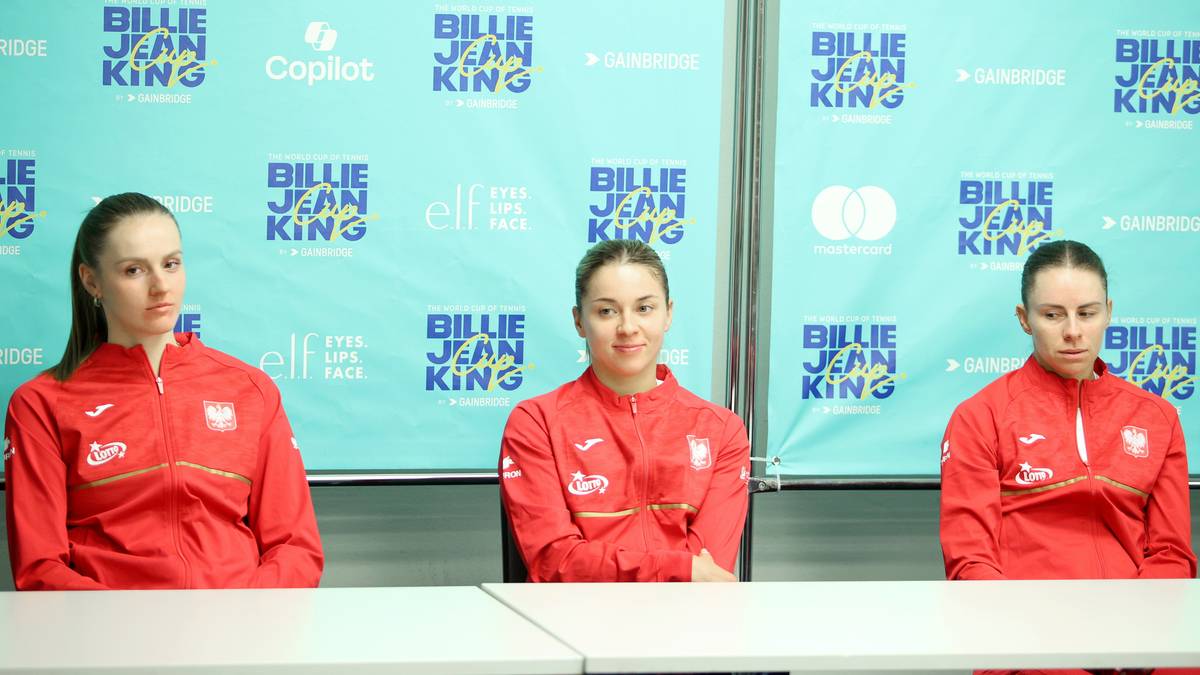 Trzy tenisistki siedzące przy stole na tle ściany z logo Billie Jean King Cup.