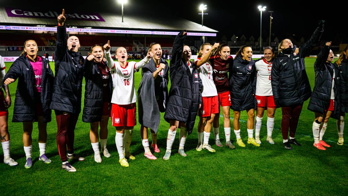 Piłkarki świętujące na boisku, niektóre z podniesionymi rękami, w białych strojach piłkarskich.