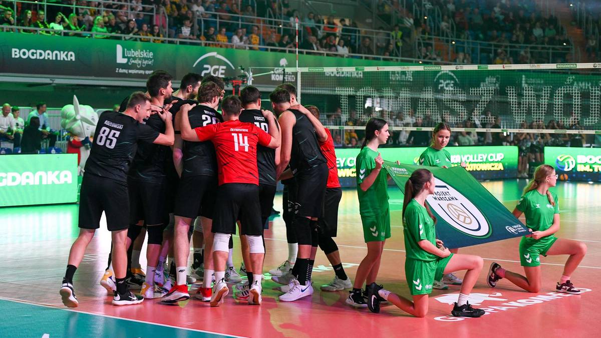 Drużyna siatkarska w strojach sportowych stoi wokół zielono-białej tablicy z logo, celebrując zwycięstwo.