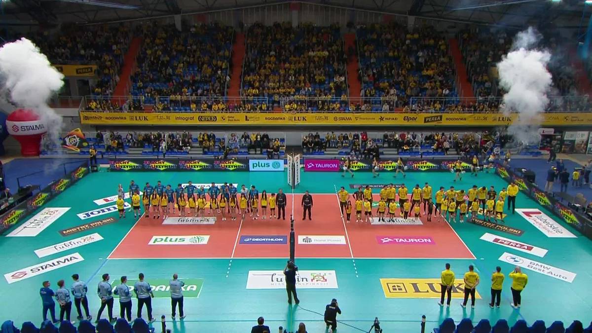 Dwie drużyny siatkarskie stoją na boisku hali sportowej przed meczem, podczas gdy widownia siedzi na trybunach.