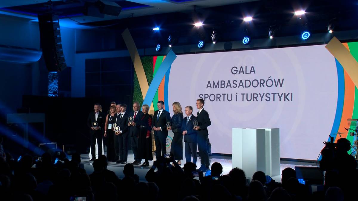 Grupa osób w strojach galowych stoi na scenie obok dużego ekranu z napisem "GALA AMBASADORÓW SPORTU I TURYSTYKI".