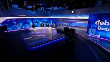 Studio telewizyjne z kilkoma osobami siedzącymi przy okrągłym stole, otoczonym niebieskimi światłami. Na ekranie w tle widoczny napis "debaty".