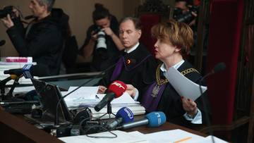 Sędzia w todze podczas rozprawy sądowej, z dokumentami w ręku i mikrofonami przed sobą.