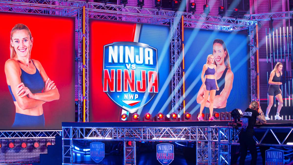 Zdjęcie przedstawia scenę z programu "Ninja vs Ninja" z widocznymi ekranami z logotypem programu i zdjęciami zawodniczek, a w tle fragment toru przeszkód.