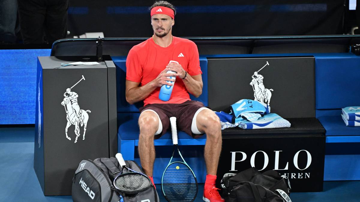 Tenisista w czerwonej koszulce i spodenkach siedzi na ławce, trzymając butelkę z wodą. Obok niego leżą torby tenisowe i sprzęt z logo "Polo Ralph Lauren".