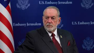 Starszy mężczyzna w garniturze stoi obok flagi USA na tle niebieskiego banera z napisem "U.S. Embassy Jerusalem".