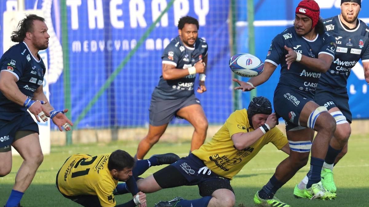 Drużyna graczy w niebieskich strojach gra w rugby z drużyną w ż&oacute;łtych strojach. Jeden z graczy w ż&oacute;łtym stroju leży na ziemi, podczas gdy inny gracz w niebieskim stroju pr&oacute;buje go podnieść.