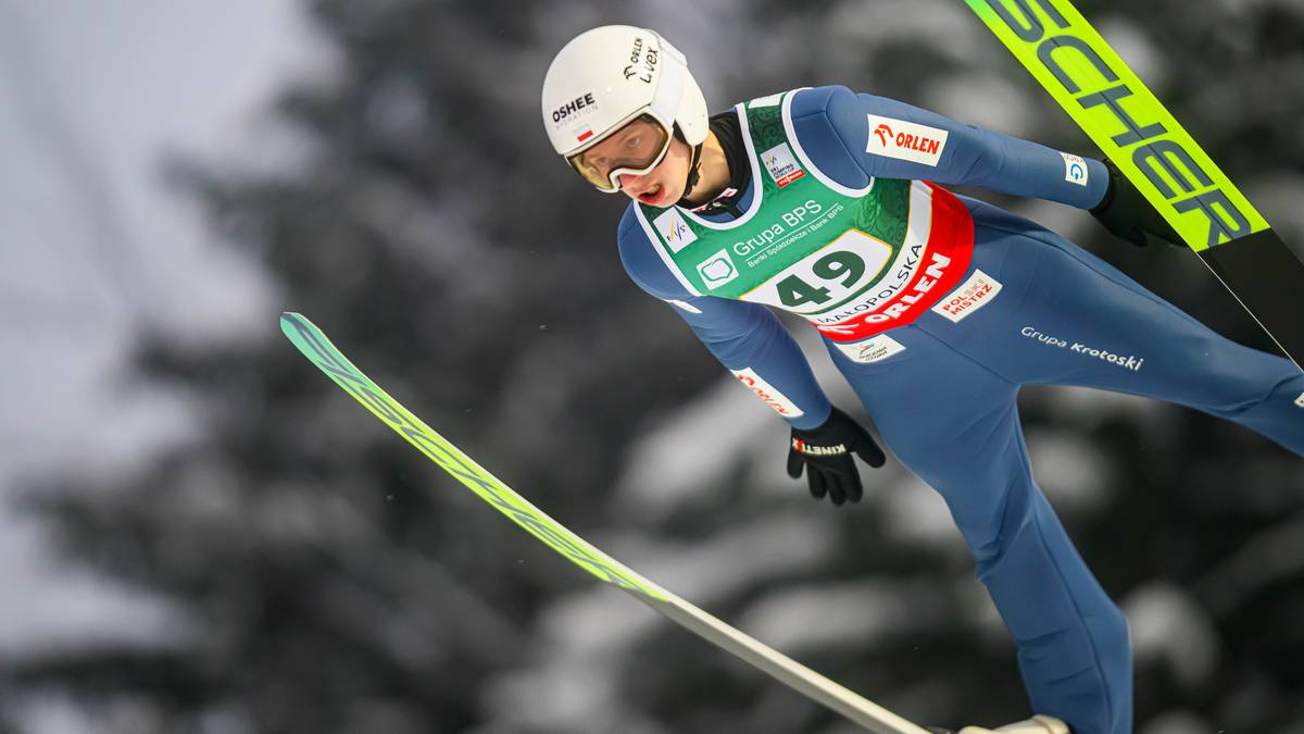 Skoczek narciarski w niebieskim kombinezonie i białym kasku wykonuje skok, z nartami skierowanymi w dół.