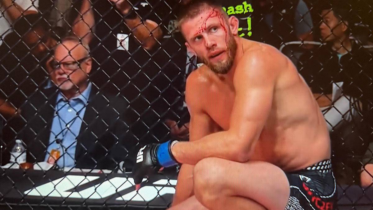 Zawodnik MMA z krwią na twarzy siedzi w klatce po walce.