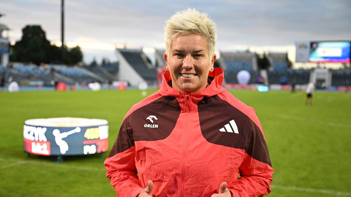 Kobieta z krótkimi blond włosami, w czerwonej kurtce sportowej, uśmiecha się do aparatu. W tle boisko piłkarskie ze stadionem.