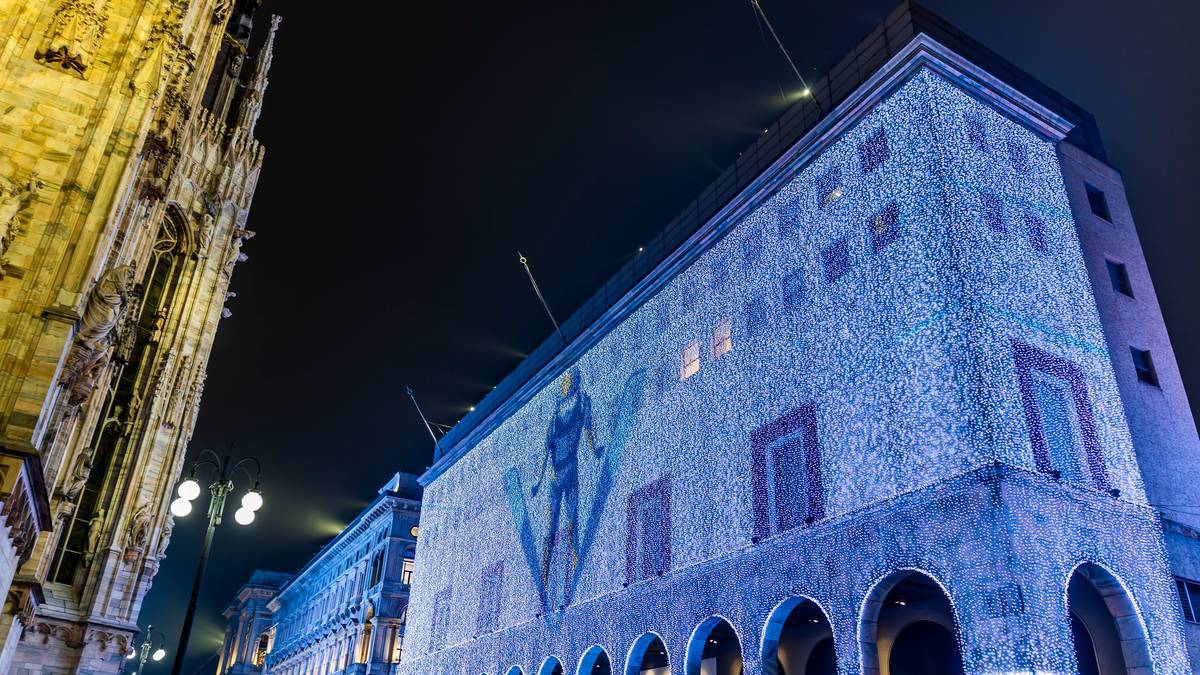 Budynek oświetlony niebieskimi światłami z projekcją postaci hokeisty na fasadzie nocą, obok zabytkowa budowla.