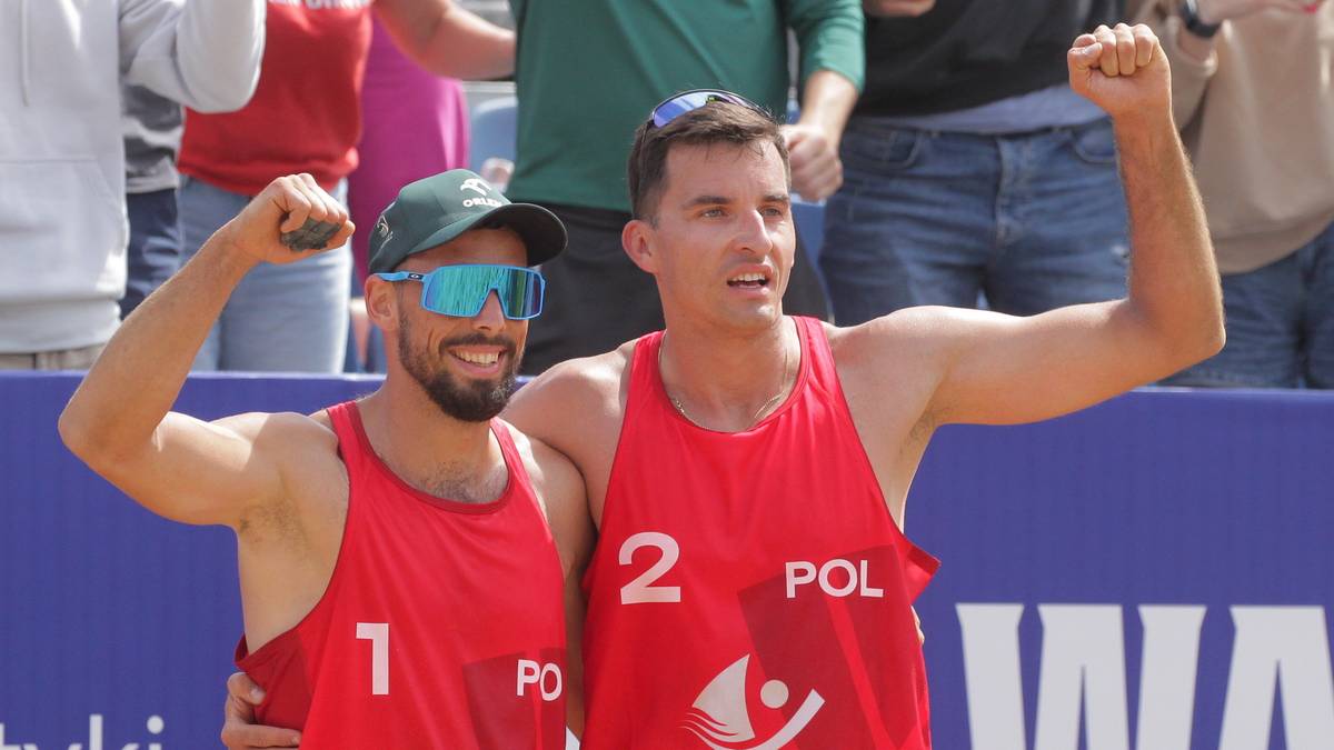 Dwóch mężczyzn w czerwonych koszulkach z numerami 1 i 2 oraz napisem "POL" na piersiach, pozuje do zdjęcia z podniesionymi pięściami. Jeden z nich ma okulary przeciwsłoneczne.