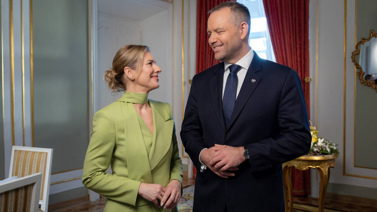 Kobieta w jasnej zielonej sukience i mężczyzna w granatowym garniturze stoją i rozmawiają w eleganckim wnętrzu.