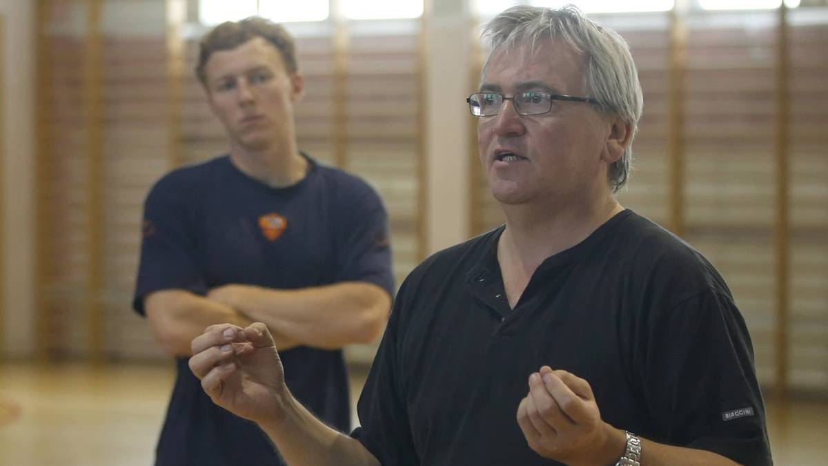 Dwóch mężczyzn w sali gimnastycznej. Na pierwszym planie starszy mężczyzna w okularach, ubrany na czarno, gestykuluje rękami. W tle młodszy mężczyzna w granatowej koszulce z emblematem.