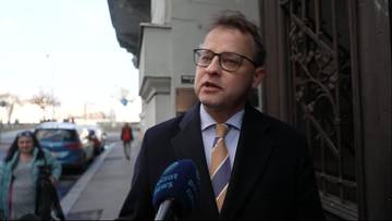 Mężczyzna w okularach i garniturze przemawia do mikrofonu.