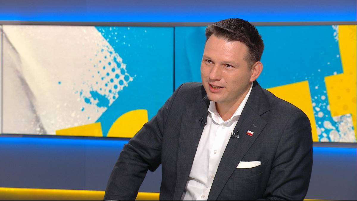 Mężczyzna w ciemnym garniturze i białej koszuli wypowiada się w studiu telewizyjnym na tle niebiesko-ż&oacute;łtej scenografii.
