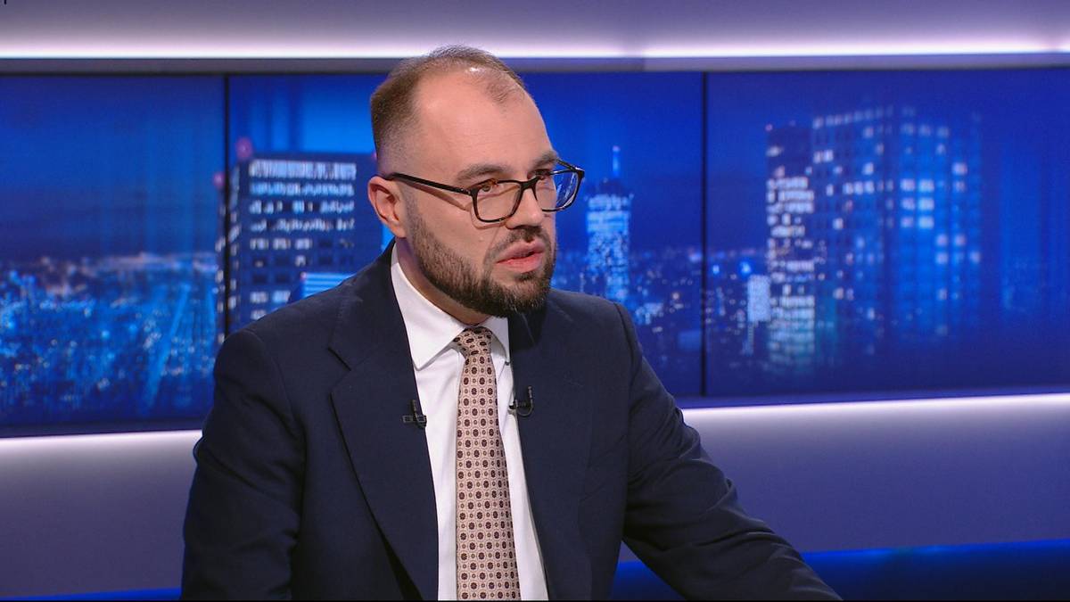 Mężczyzna w okularach i garniturze siedzi i m&oacute;wi, na tle widać fragmenty nocnego miasta.