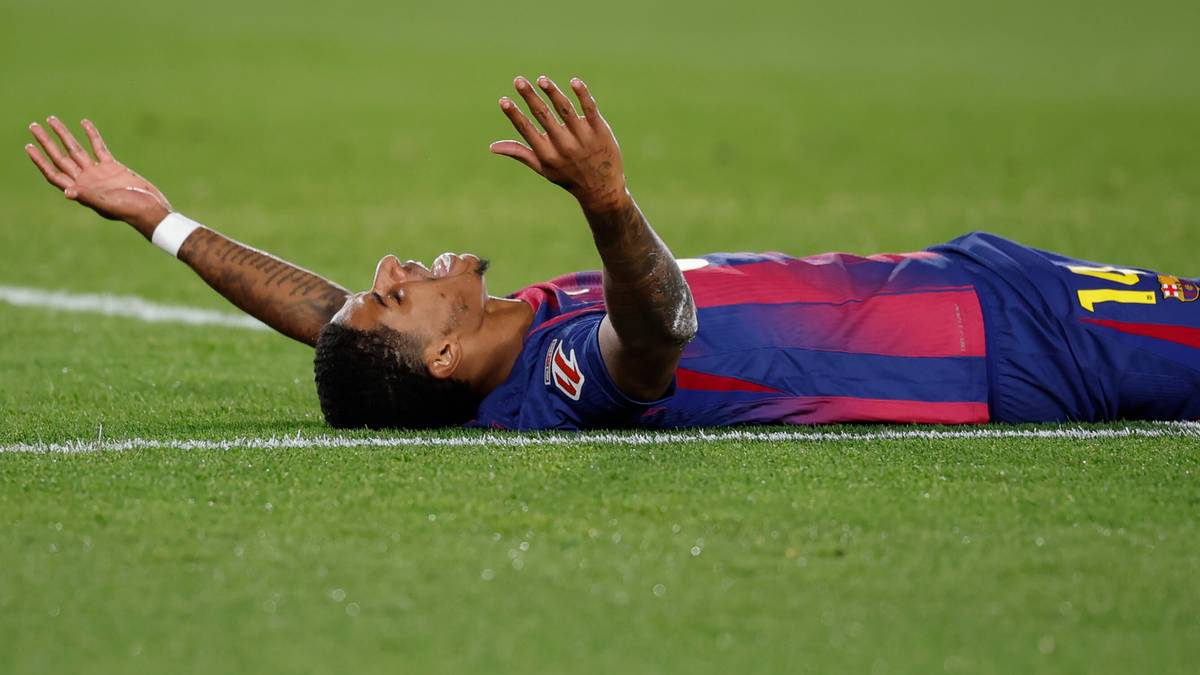 Piłkarz leżący na trawie z uniesionymi rękami, w niebiesko-bordowym stroju piłkarskim.