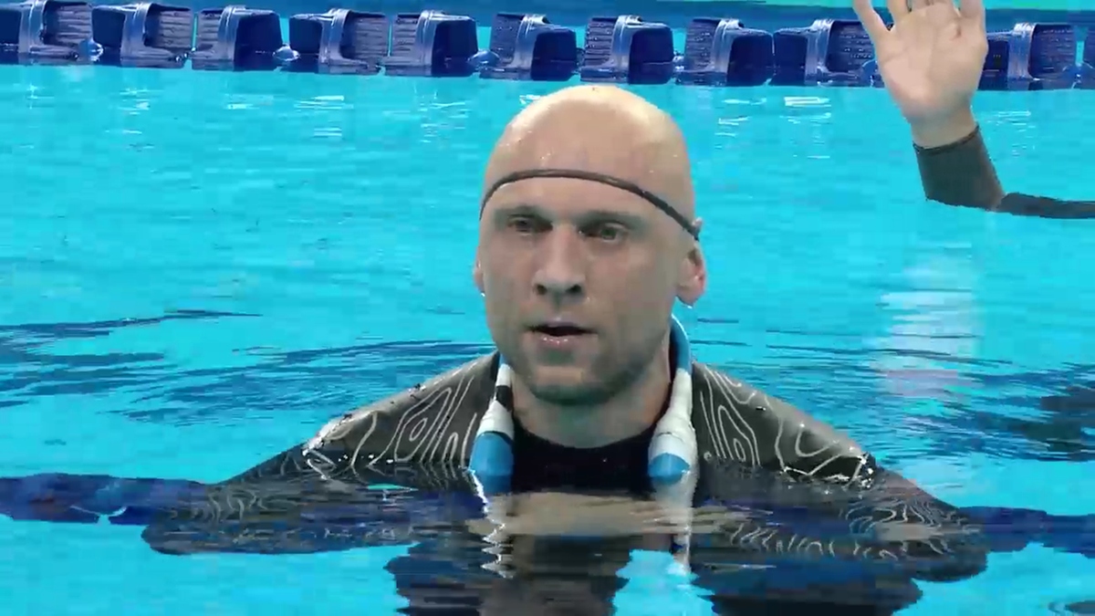 Łysy mężczyzna w stroju pływackim z czarnym wzorem i niebieskimi akcentami wystaje z basenu, patrząc przed siebie. W tle widać niebieską wodę i krawędź basenu z oznaczeniami.