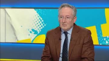 Mężczyzna w okularach i brązowej marynarce siedzi przed kolorowym tłem.