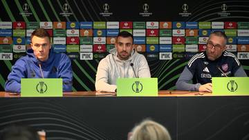 Trzech mężczyzn siedzi za stołem podczas konferencji prasowej, przed nimi stoją zielone tablice z logo.