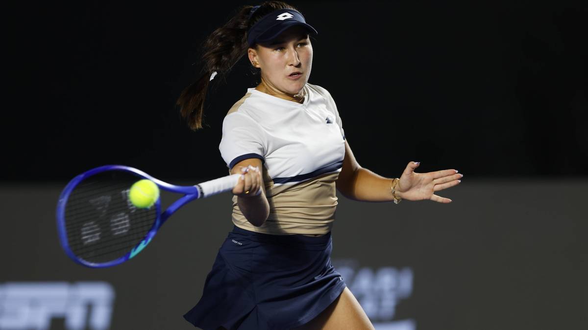 Tenisistka w białej koszulce i granatowej spódniczce uderza piłkę rakietą podczas meczu.