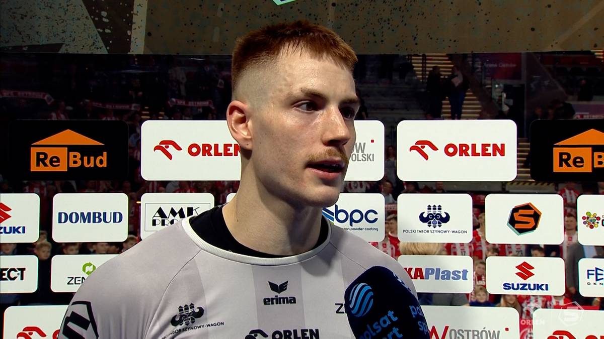 Mężczyzna w szarej koszulce sportowej z logotypem Orlen, na tle ścian z logotypami sponsorów, z mikrofonem skierowanym w jego stronę.