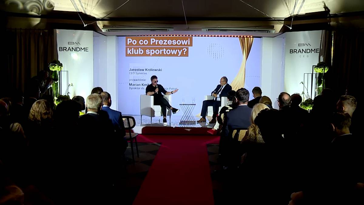 Dwóch mężczyzn siedzi na scenie na krzesłach naprzeciw siebie, przed dużym ekranem z napisem "Po co Prezesowi klub sportowy?". Jeden mężczyzna gestykuluje podczas rozmowy. Widownia siedzi w ciemnościach w tle.