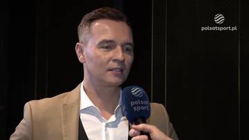 Mężczyzna w jasnej marynarce i białej koszuli, trzymający mikrofon z logo "Polsat Sport".