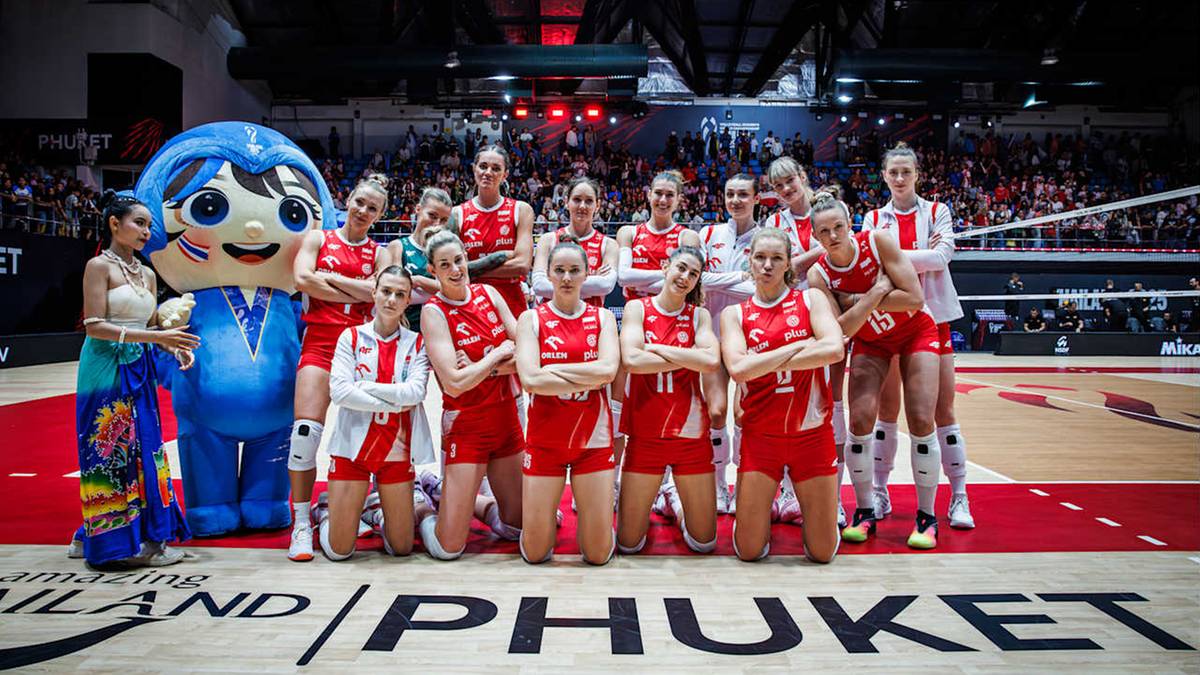 Grupa siatkarek w czerwonych strojach stoi na boisku siatkarskim obok niebieskiej maskotki. Na widowni widać publiczność.