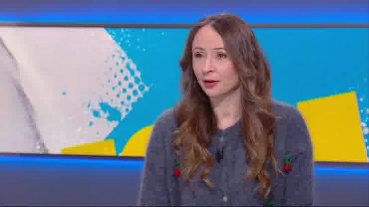 Kobieta z długimi brązowymi włosami w szarym swetrze z haftami, siedząca przed kolorowym tłem.