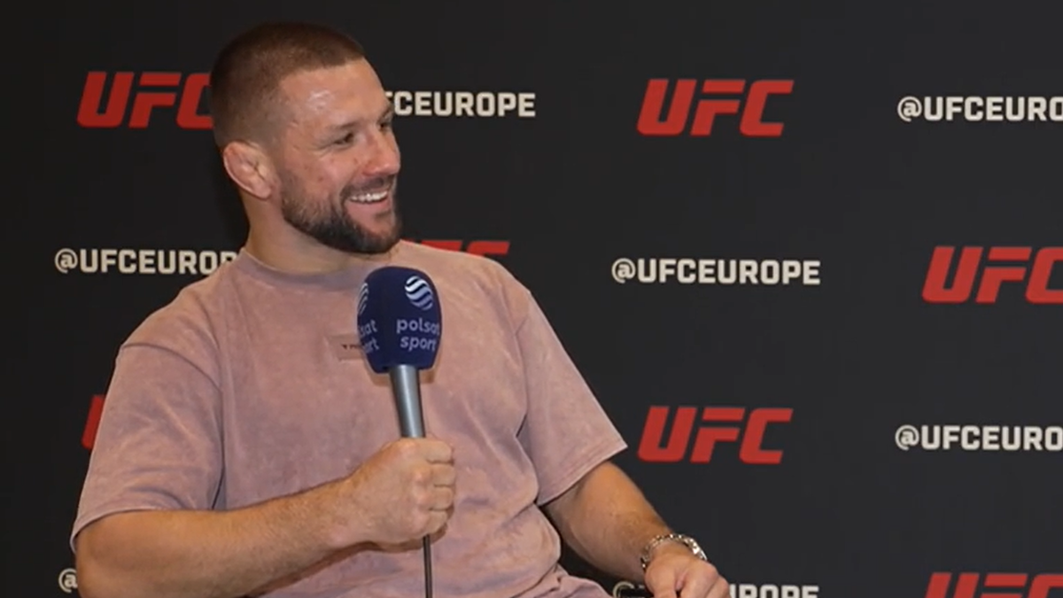 Szczęśliwy mężczyzna w różowej koszulce siedzi i trzyma mikrofon z logo "Polsat Sport". W tle widnieje czarne logo UFC.