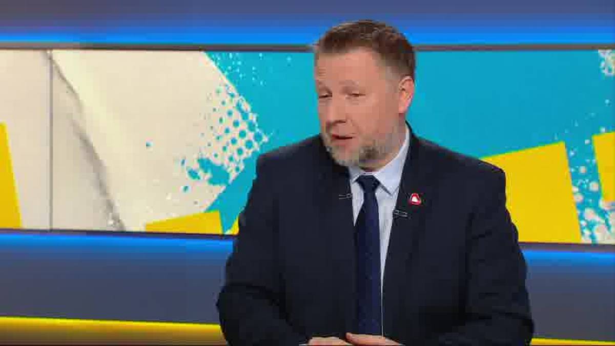 Mężczyzna w garniturze i krawacie siedzi przed ekranem z kolorowym wzorem graficznym.