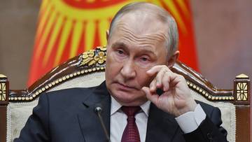 Władimir Putin siedzi na krześle, w garniturze i krawacie, z ręką przy twarzy.